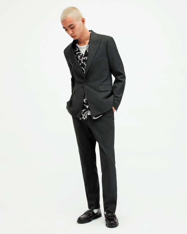 Photograph showing a male model wearing a black blazer, black trousers and a printed black shirt with black leather loafers.