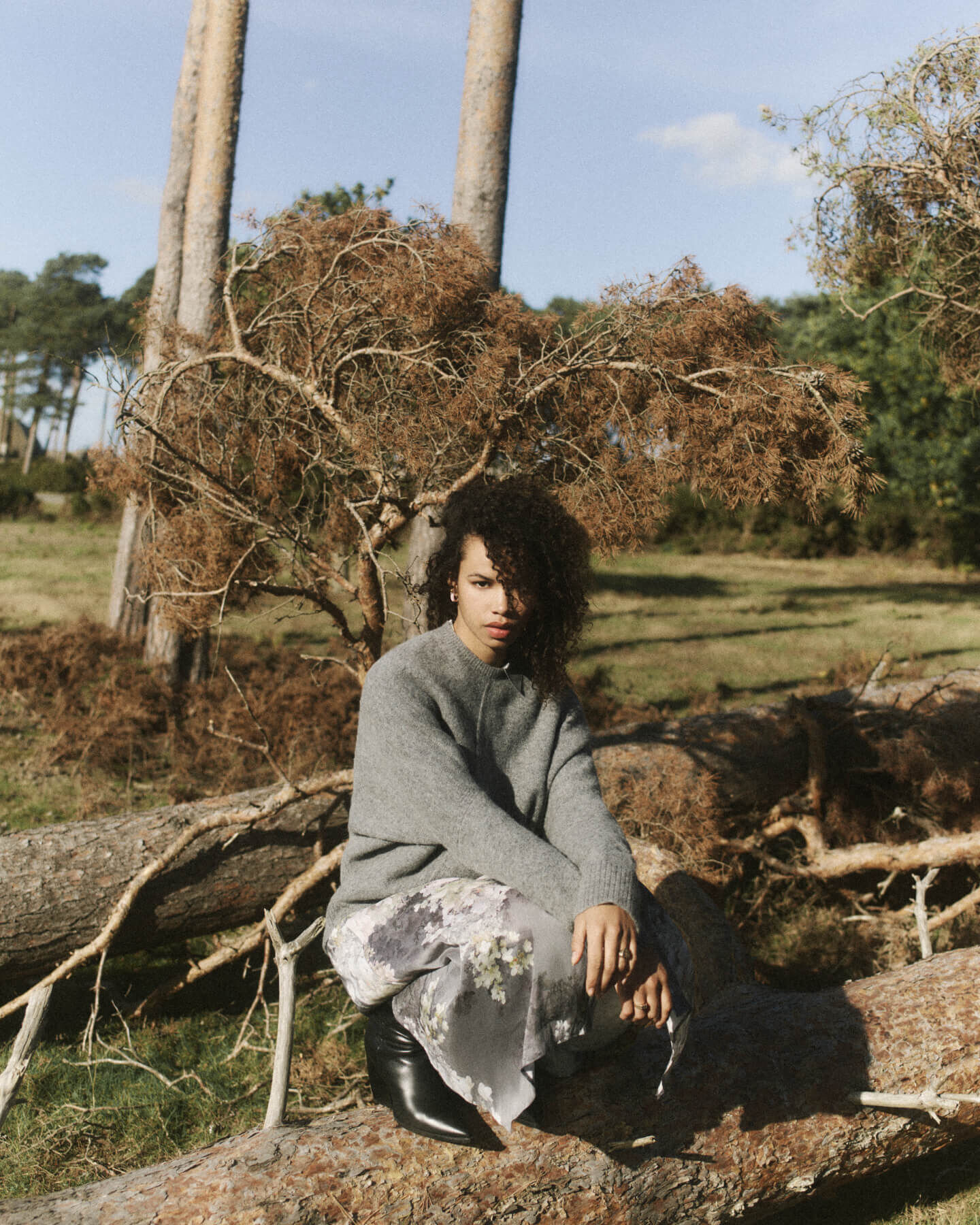 Woman in a grey jumper and dress with black leather boots crouching on a fallen tree