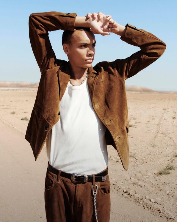 Un homme se tenant dans le désert portant un blazer marron clair en cuir suédé  et un collier en argent sterling, avec un pantalon marron assorti, une ceinture en cuir et une chaîne de ceinture. 