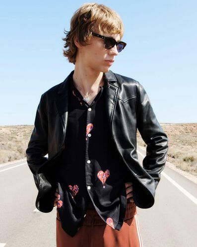 Man wearing a black leather jacket over a black heart print shirt and red trousers