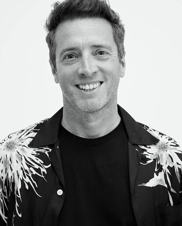 Black and white headshot of a male wearing a black top with a Hawaiian shirt layered in front on a white background 