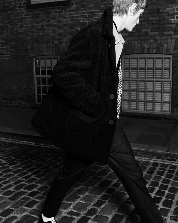 A black and white photograph of a man wearing a long black coat and black trousers walking on paved ground.