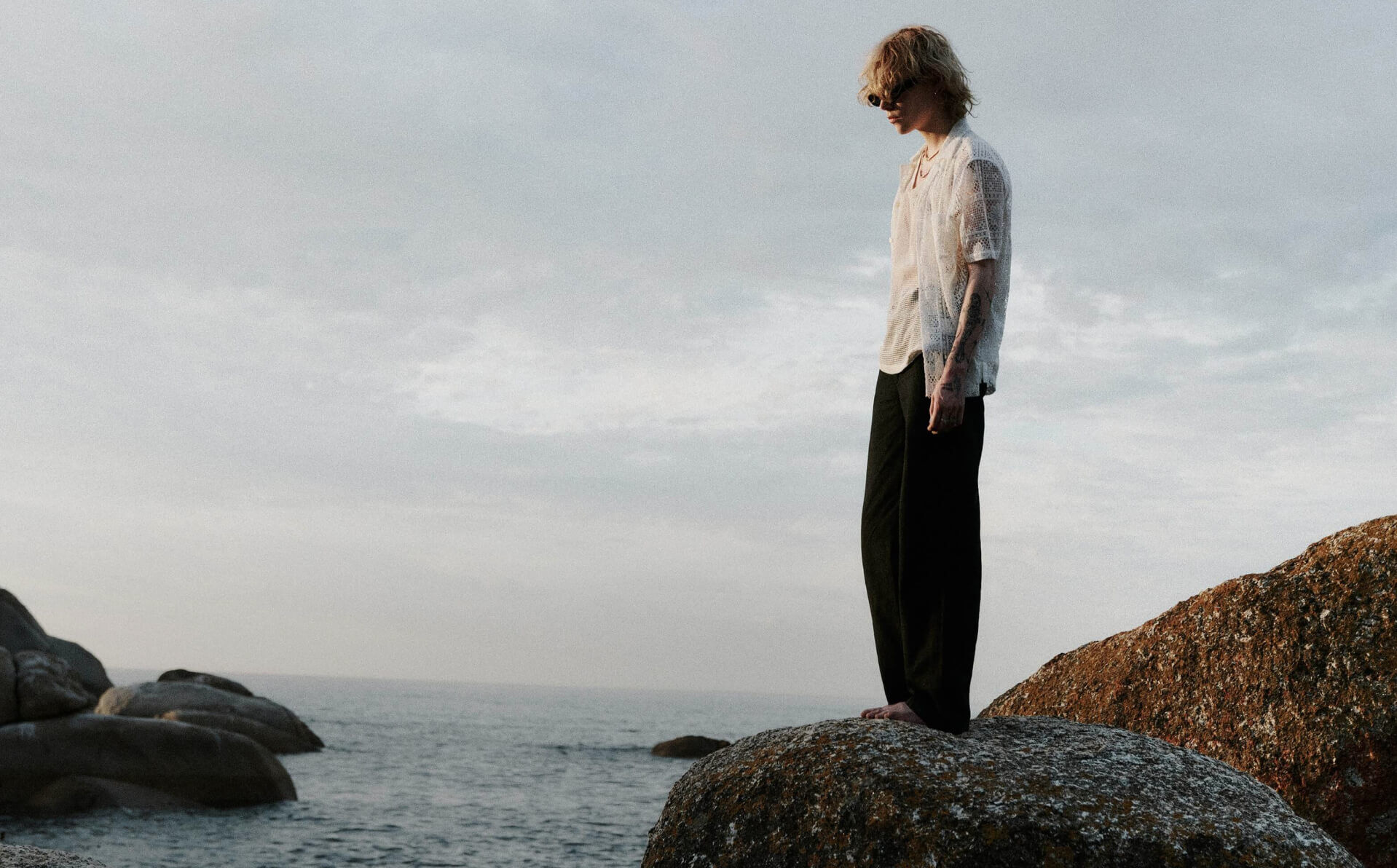Herr mit weißem Hemd und schwarzer Hose steht auf einem Felsen am Meer