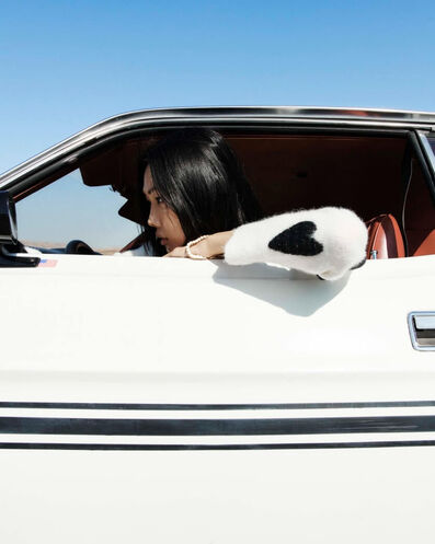 Une mannequin assise dans une voiture ancienne blanche, avec le bras accoudé sur le rebord de la portière montrant un cœur noir sur le cardigan.