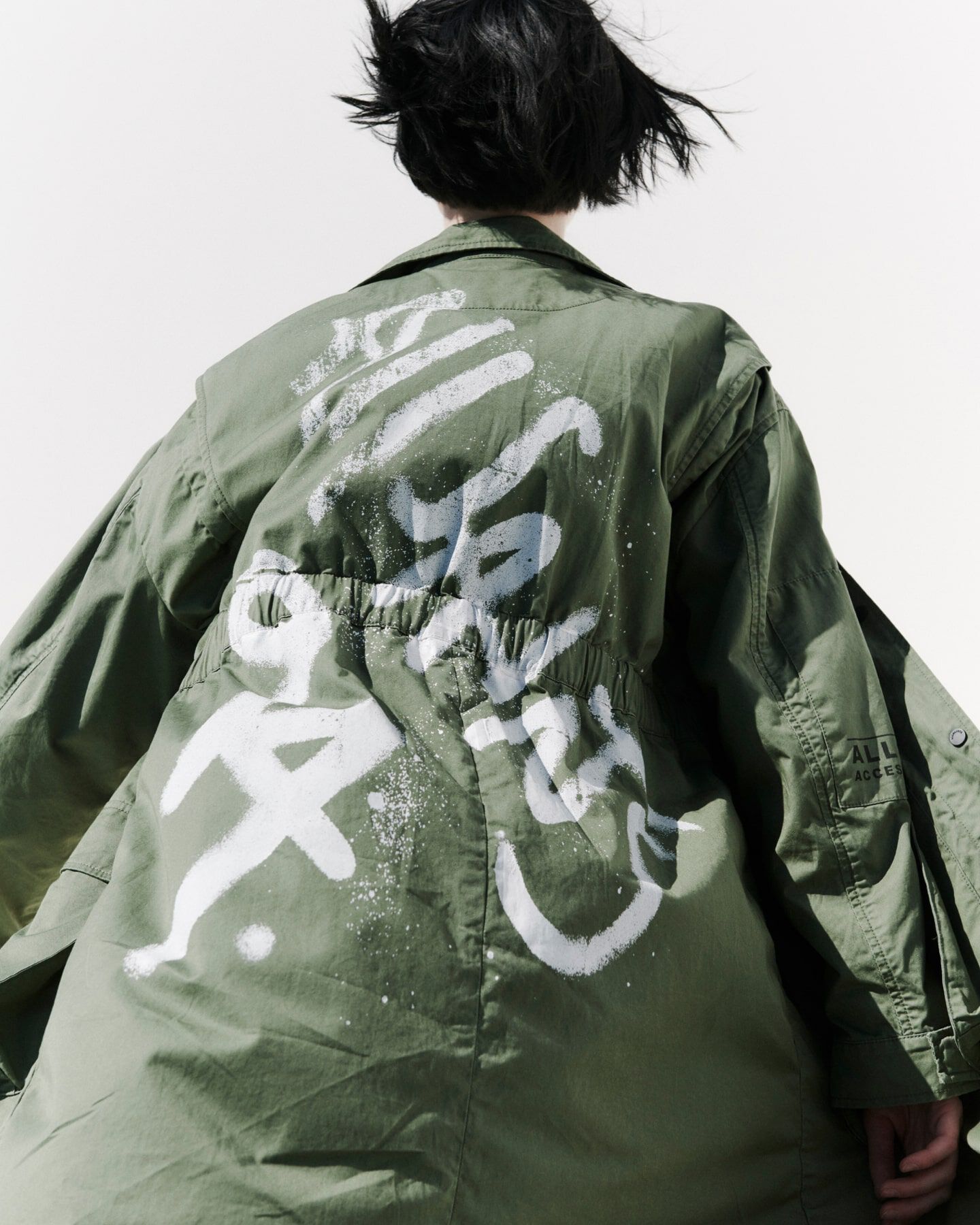 A woman wearing a light green jacket with AllSaints 94 spray painted on it in white