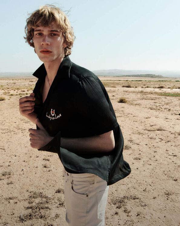 Man wearing a black Hawaiian shirt with embroidery and with white jeans walking in the desert.
