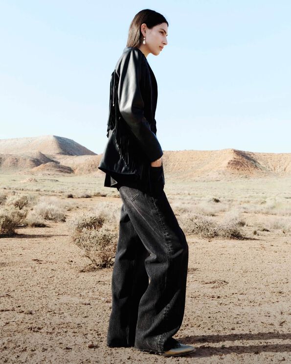 Ein Profilfoto eines Models, das eine schwarze Lederjacke mit Fransen und schwarze Jeans mit weitem Bein trägt und in der Wüste steht.