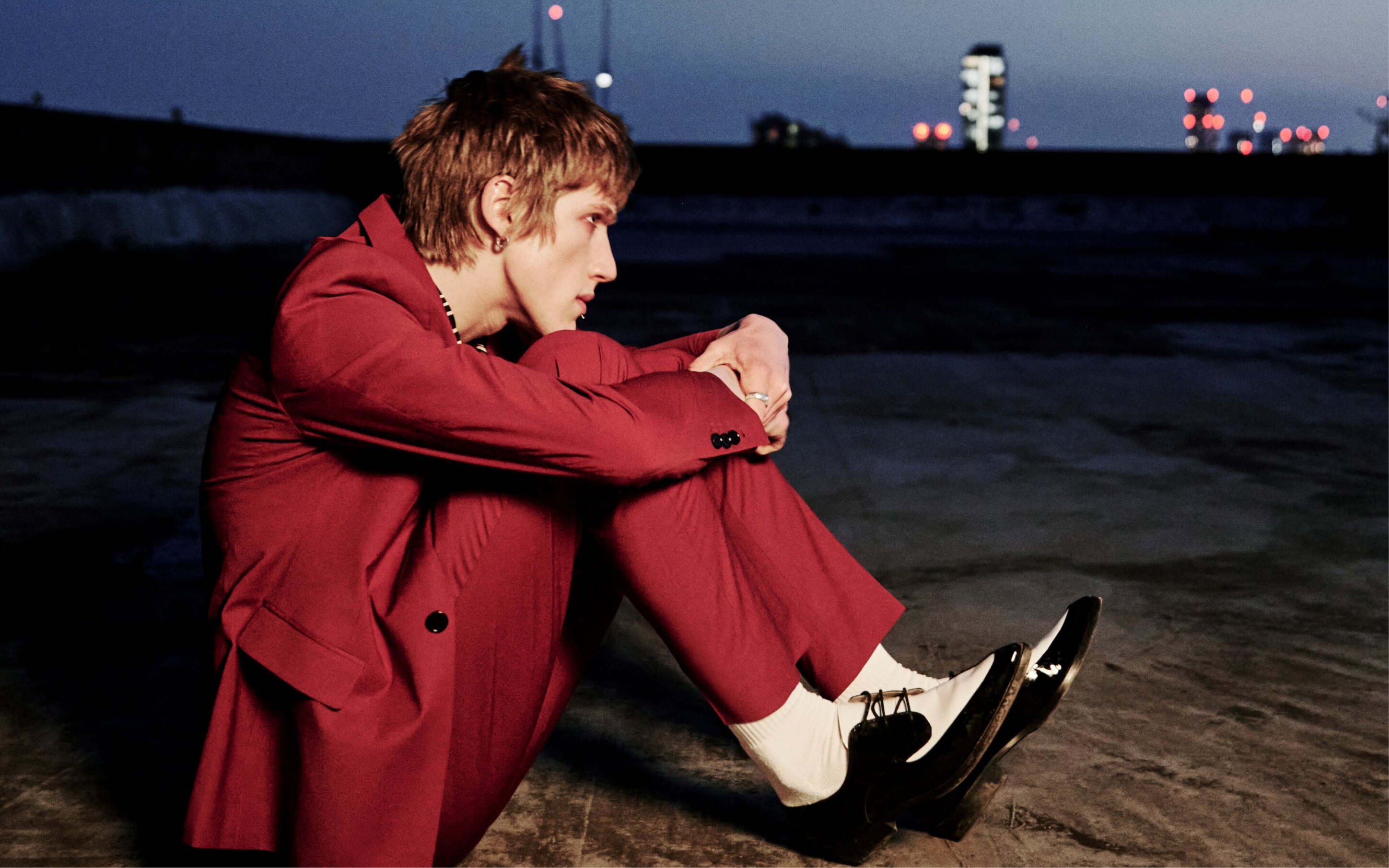 Man wearing a red tailored suit and black and white dress shoes sitting on the beach at nighttime.