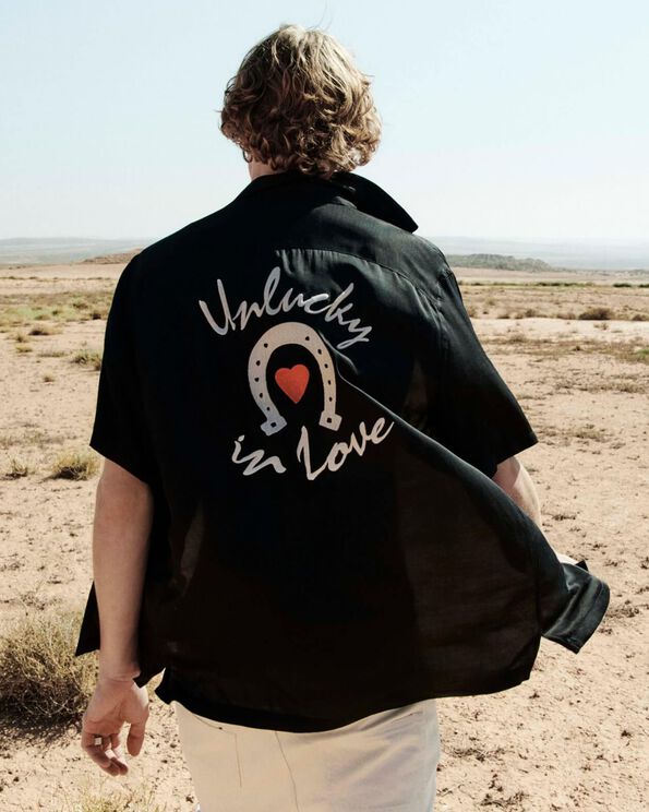 Photon dun homme se tenant de dos dans le désert et portant une chemise noire brodée avec un jean blanc.