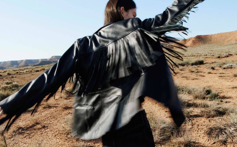 Photograph showing a model dancing while wearing a fringed black leather jacket in the desert.