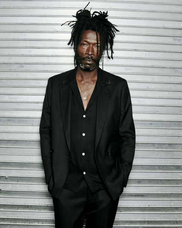 A man with dreadlocks wearing a black blazer, shirt and trousers standing with his hands in his pockets.