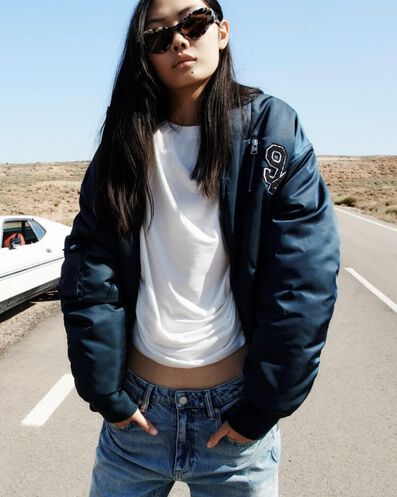 Model trägt eine dunkelblaue College-Jacke, ein kurzes weißes T-Shirt, Jeans mit niedriger Taille und eine Sonnenbrille und steht auf einer Straße in der Wüste.