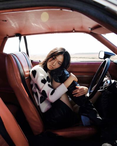 Foto eines Models, das in einem altmodischen Auto sitzt und eine weiße Strickjacke mit schwarzen Overknee-Lederstiefeln trägt.