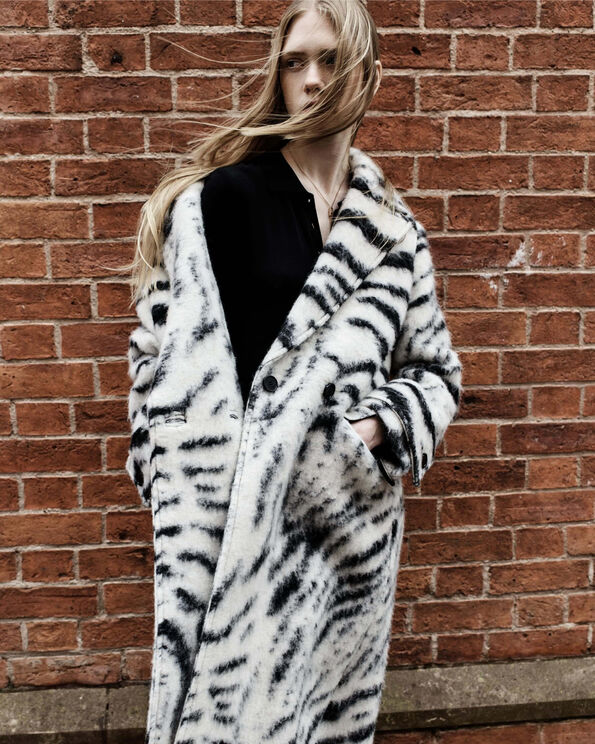 A woman with blonde hair swaying on the wind wearing a zebra-print coat standing in front of a bricked wall.