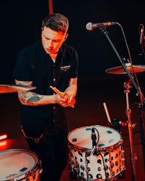 Drummer plays the drums and wears a black polo shirt and black pants.