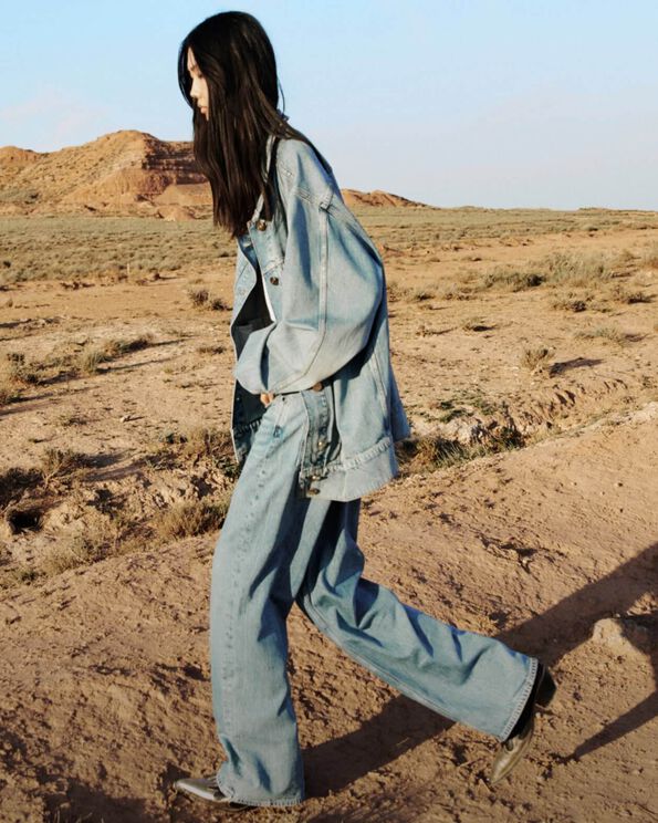 A model wearing a denim jacket and denim jeans with leather cowboy boots walking in the desert.