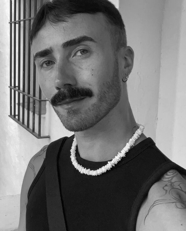 A black and white headshot of a man with a moustache wearing a black tank and a white beaded necklace