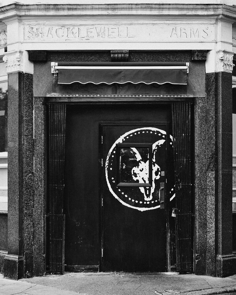 Black-and-white photograph of a ram standing beside a wooden farm gate, with the AllSaints ram skull logo overlaid above.