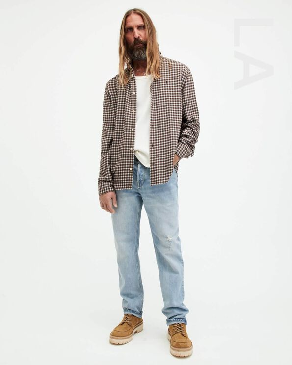 Portrait of a male model wearing a checked shirt over a white t-shirt with light blue jeans and brown suede shoes with LA written on the right hand side.