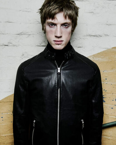 A man wearing a black leather jacket with the zip fastened standing in front of a white and yellow wall.
