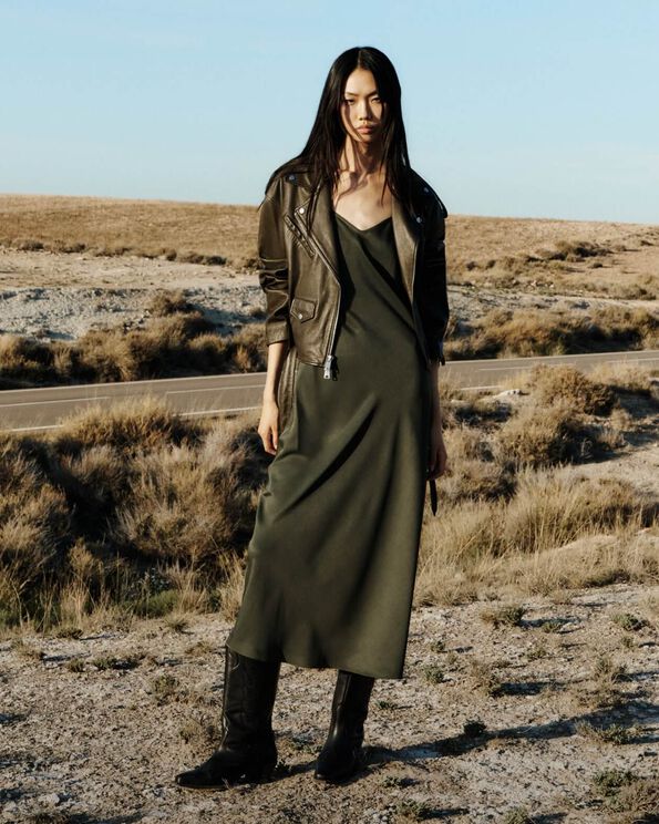 Model trägt eine dunkelgrüne Lederjacke über einem khakifarbenen Unterkleid und Cowboystiefel und steht in der Wüste.