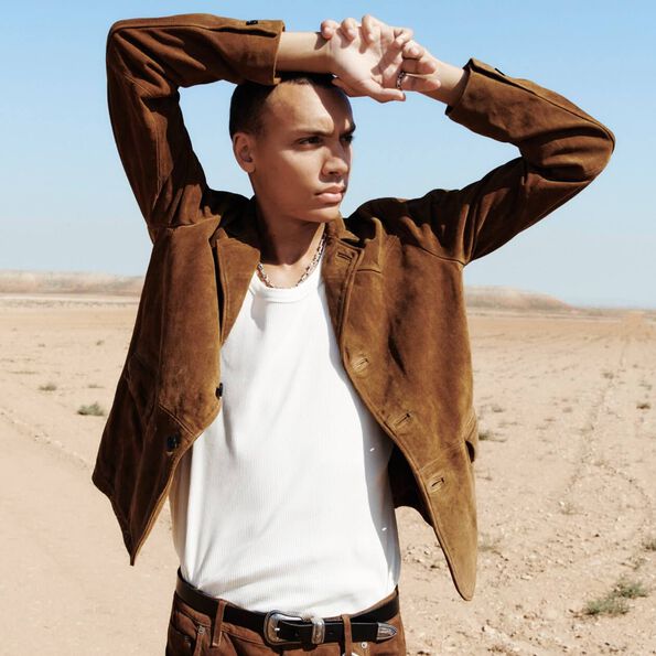 A man in the desert wearing a brown leather jacket with a white top and trousers