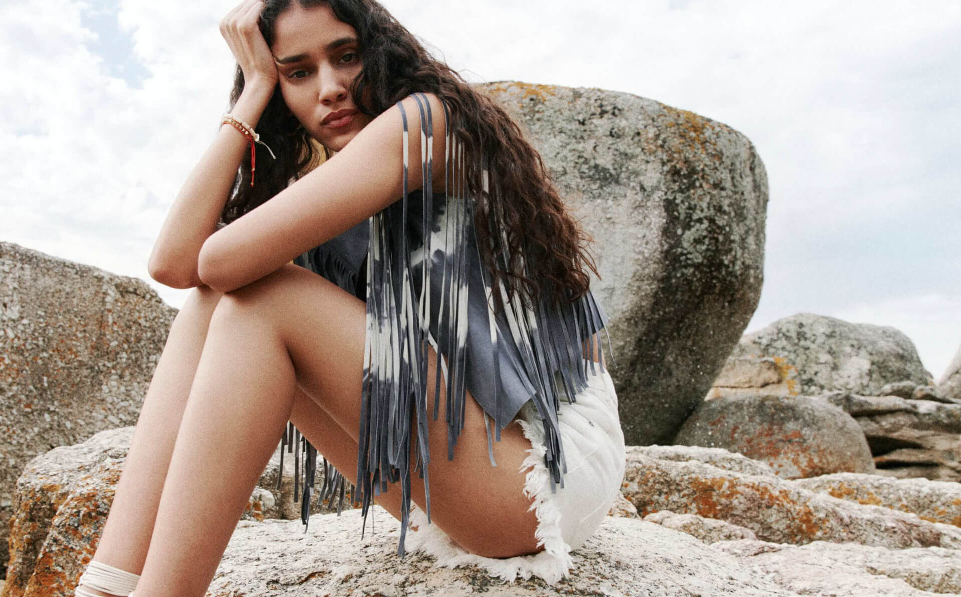 Woman wearing a grey tie dye fringed waistcoat and white shorts sat on a boulder 