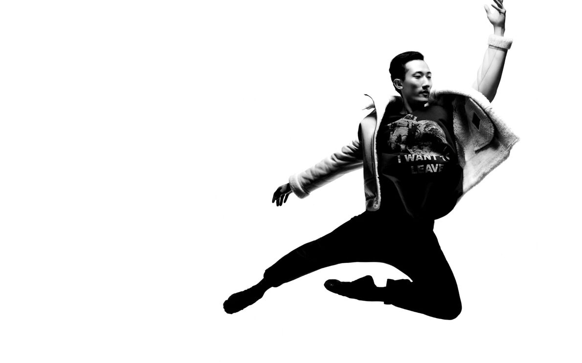 A black and white photograph of a man wearing a shearling over a black graphic t-shirt leaping.