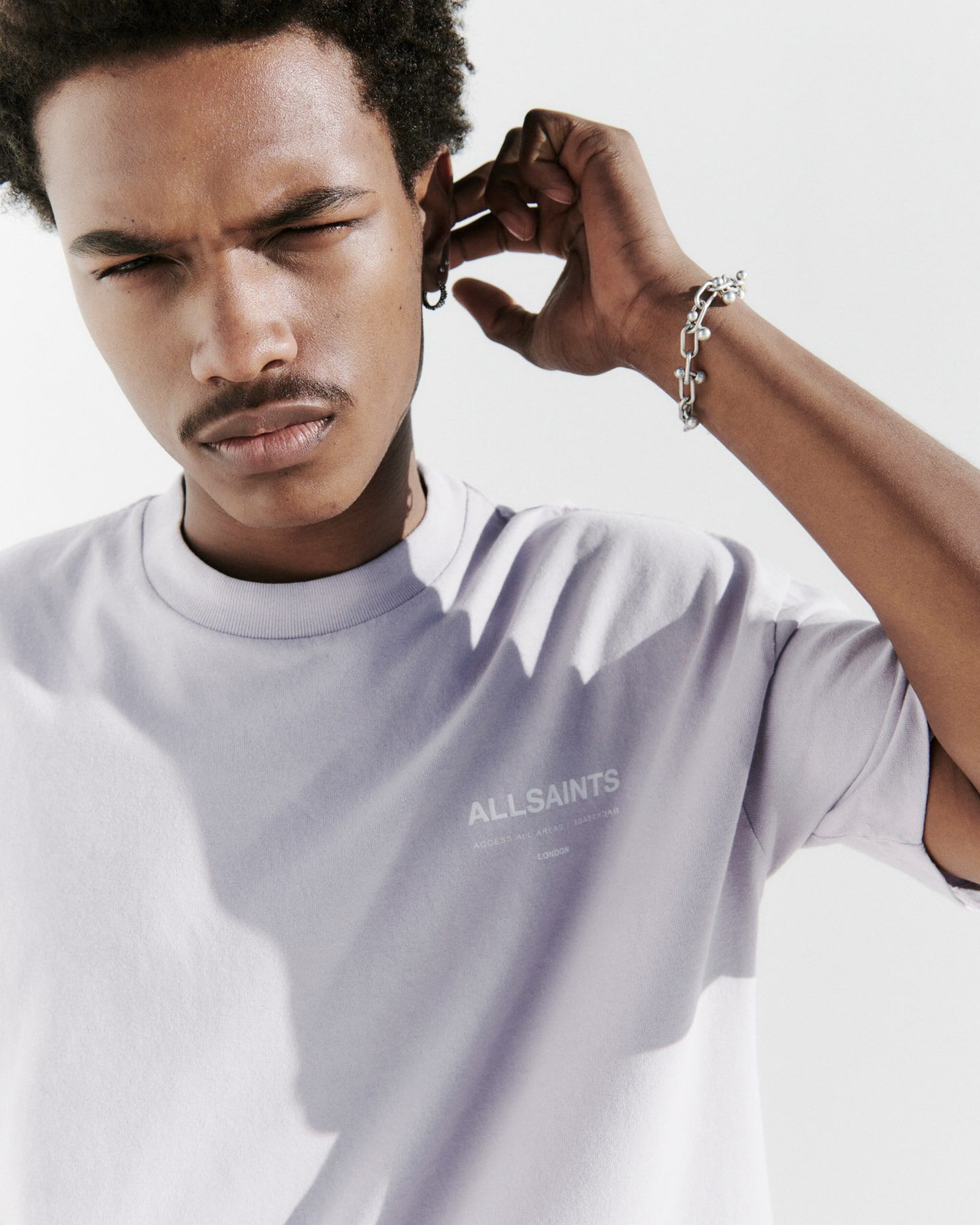 A man wearing a lilac t-shirt with AllSaints logo and a chain sterling silver bracelet touching his ear
