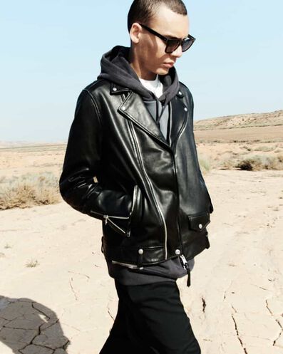 Man wearing a black leather jacket with a black hoodie, sunglasses and pants in the desert.