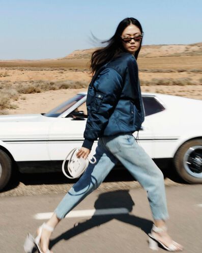 Model wearing a dark blue varsity jacket and light blue jeans with heeled sandals, walking and holding a white leather handbag.