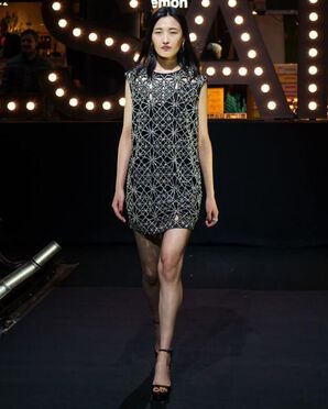 A woman wearing a black mini dress embellished with silver sparkly details and black heeled shoes catwalking.
