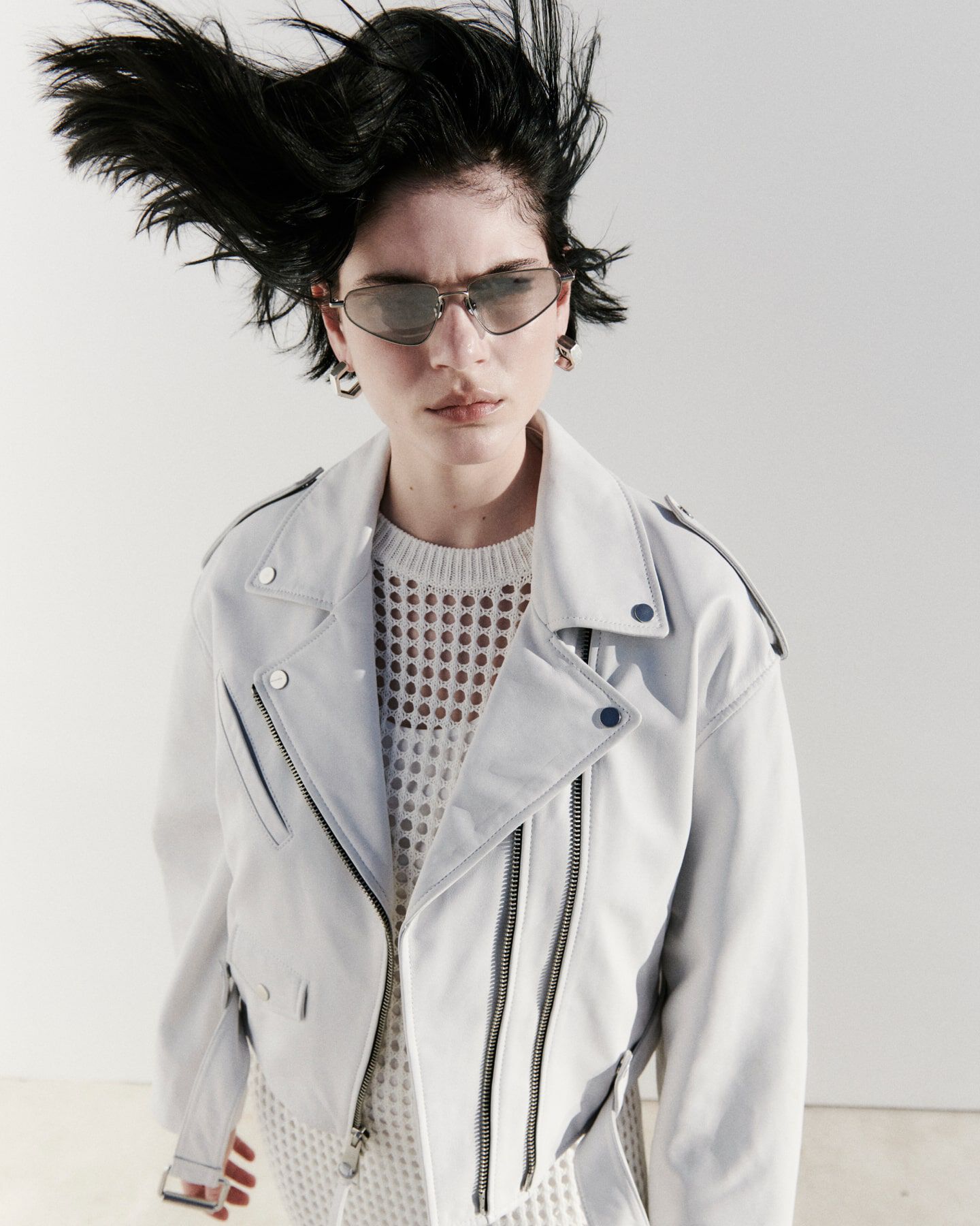 Ein weibliches Model trägt eine weiße Lederjacke, eine spitze Sonnenbrille und einen weißen Lochstoff Pullover.