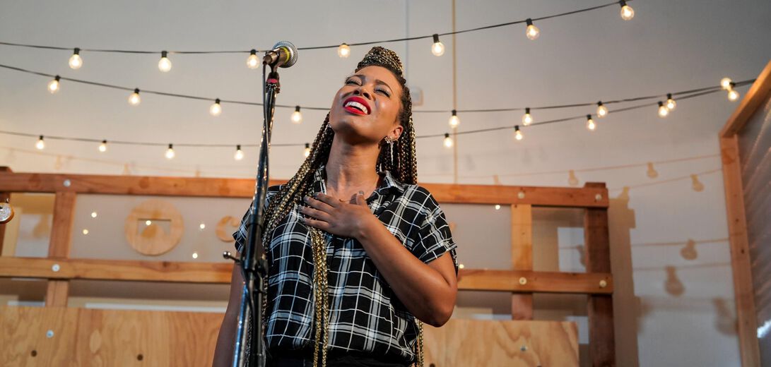 Photograph showing a female singer wearing a checked short sleeve shirt singing into a microphone, in front of a background showing string lights and wooden panels.