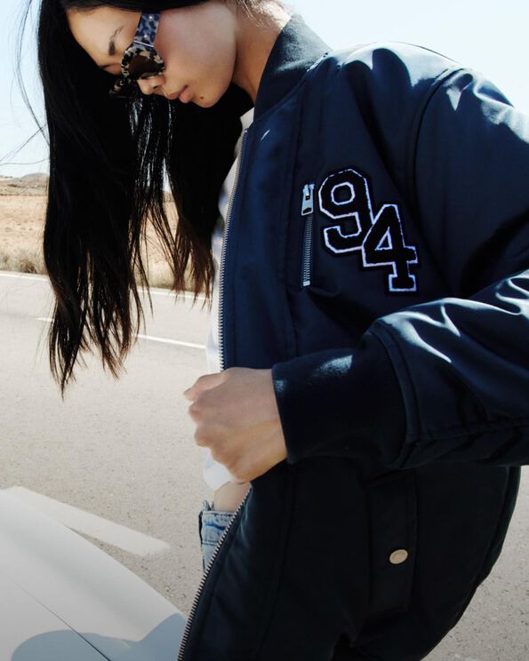 Model looking down with sunglasses and opening her dark blue varsity jacket.
