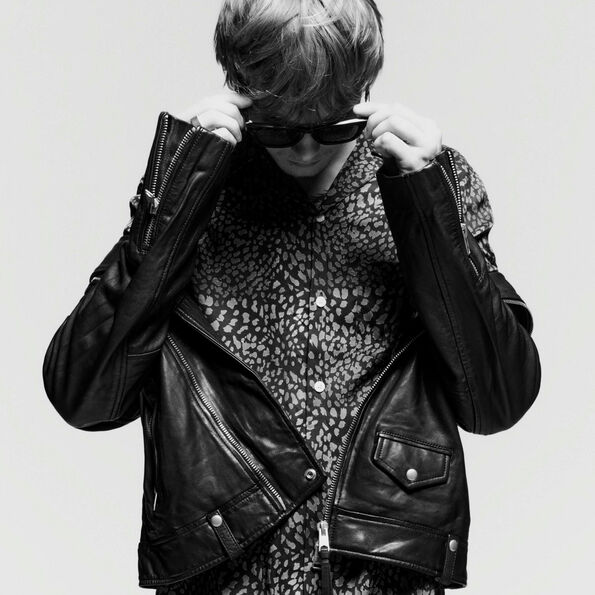 Black and white photograph of a man wearing an unzipped black leather jacket over a leopard print shirt putting sunglasses on