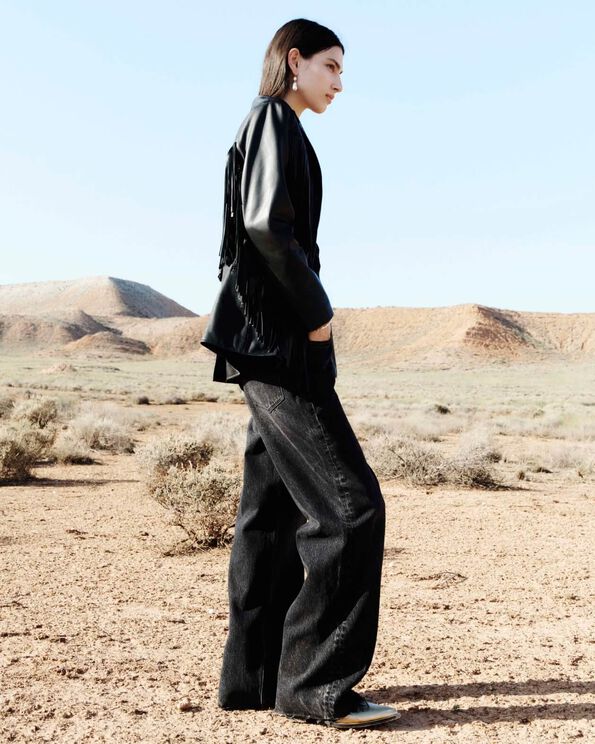 A profile photograph of a model wearing a fringed black leather jacket with black wide leg jeans standing in the desert.