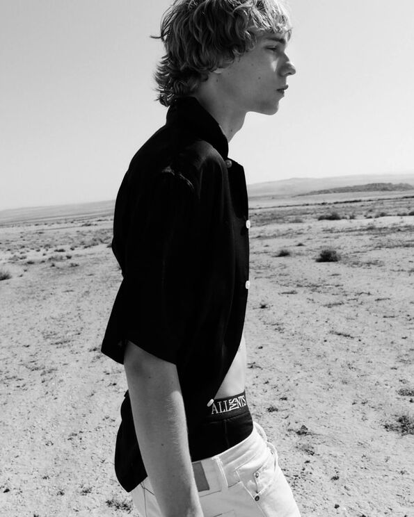 Schwarz-Weiß-Foto eines Mannes, der ein offenes schwarzes Hemd, schwarze AllSaints-Boxershorts und weiße Jeans trägt und in der Wüste spaziert.