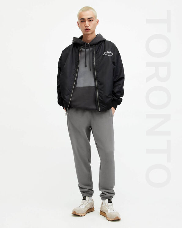 Portrait of a male model wearing a black bomber over a hoodie with grey sweatpants and white and beige trainers with TORONTO written on the right-hand side..