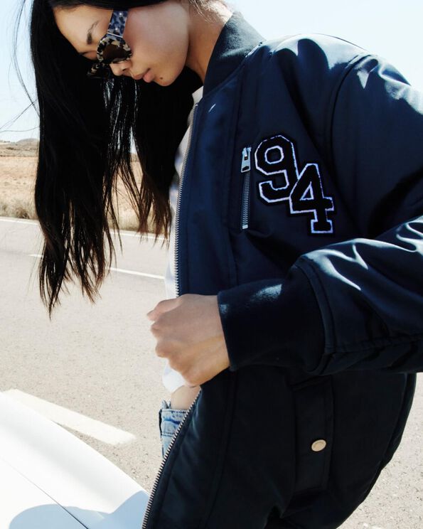 Model looking down with sunglasses and opening her dark blue varsity jacket.