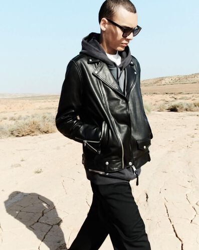 Man wearing black sunglasses, a black leather jacket over a hoodie and black pants walking in the desert.