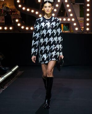 A woman wearing a short houndstooth print dress with high black boots catwalking.