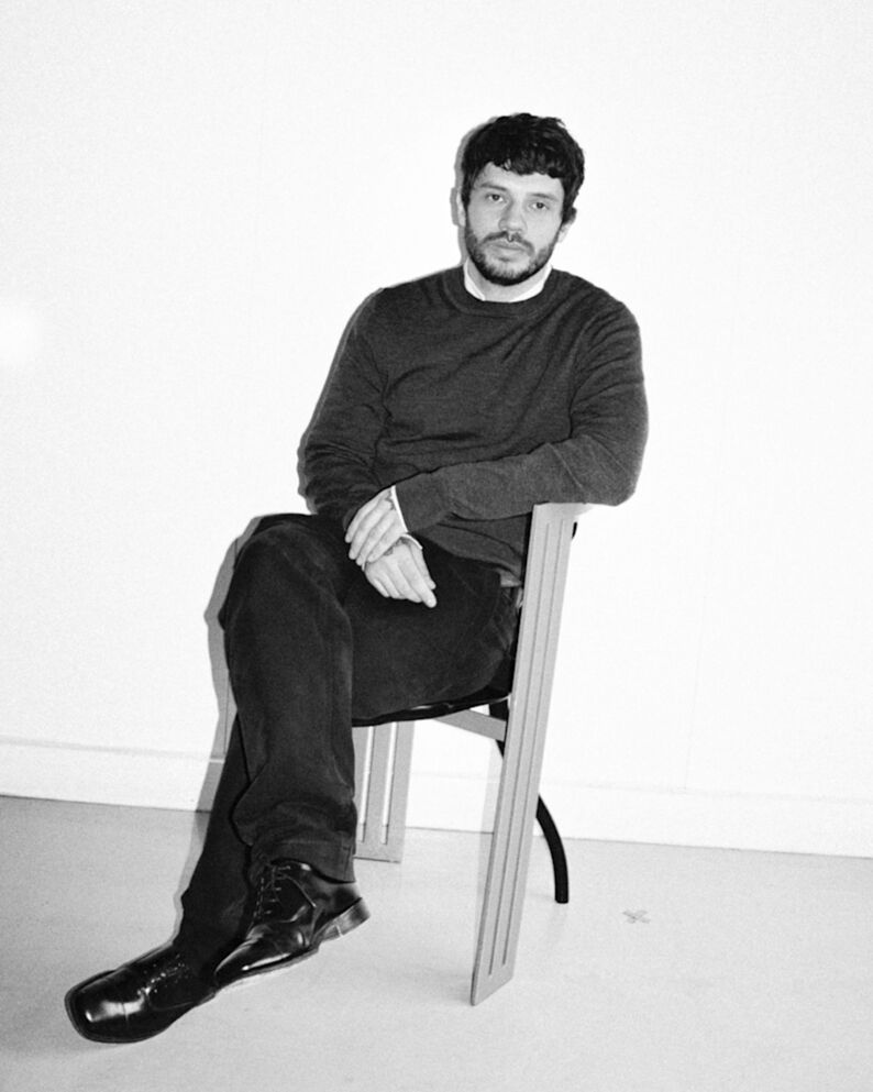 Black-and-white portrait of Aaron Esh seated cross-legged on a wooden chair, gazing calmly at the camera in a minimalist room.