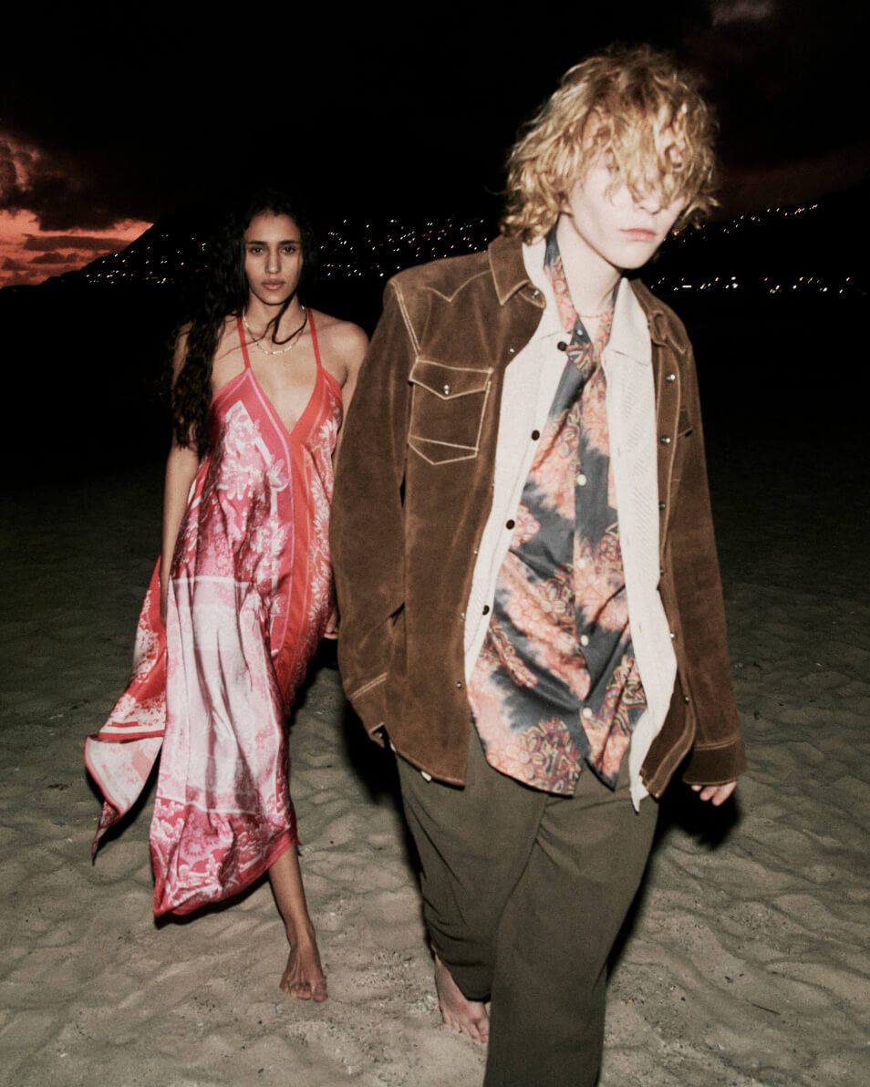 Man wearing a shirt, brown leather jacket and pants walking on a beach at night with a woman in a red dress