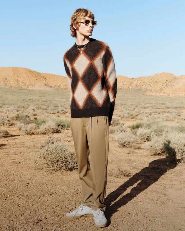 A man wearing beige sunglasses, an diamond motif brown jumper and brown trousers with white trainers in the desert.