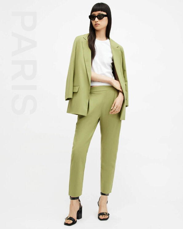Portrait of a model wearing a light green blazer and trousers with black sandals et black sunglasses with PARIS written on the left hand side.