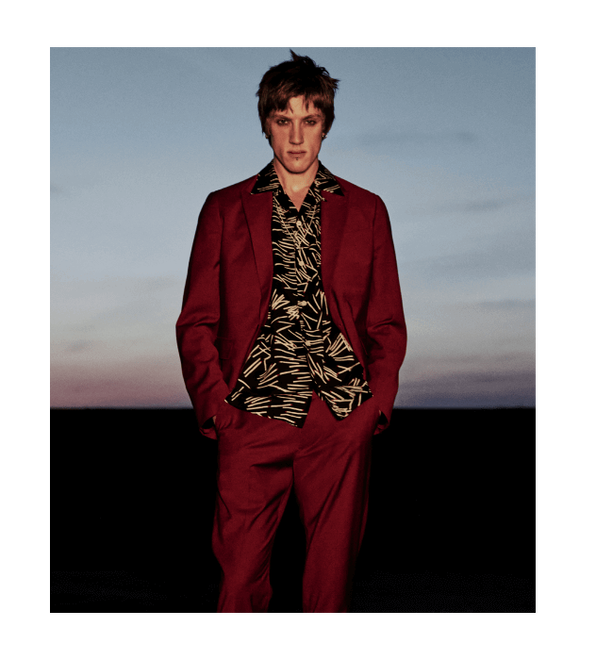 Photograph of man wearing a red suit with abstract print black and white shirt standing in front of the sunset.
