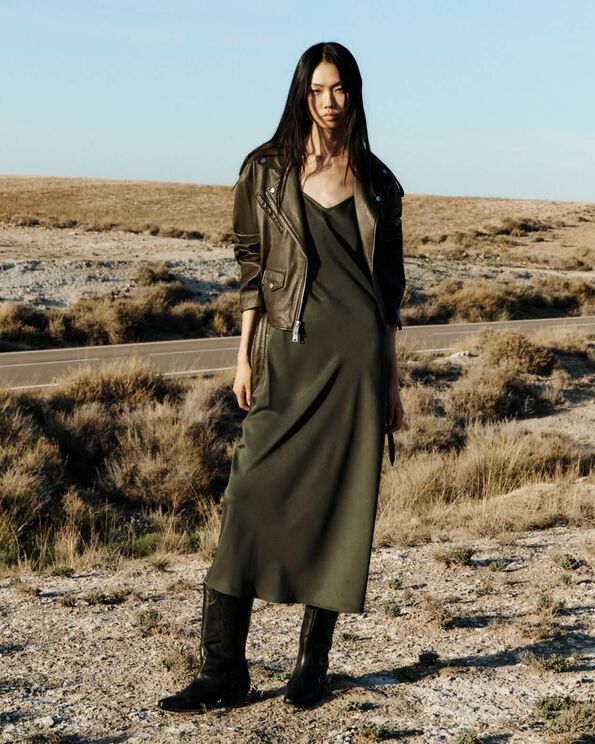 Model wearing a dark green leather jacket over a khaki slip dress and cowboy boots, standing in the desert.