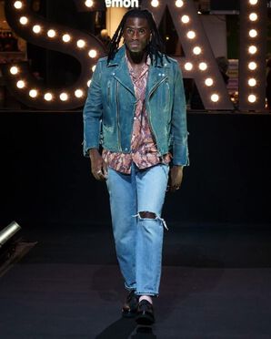 A man wearing a teal leather jacket over a snake print shirt and a pair of jeans ripped at the right knee catwalking.