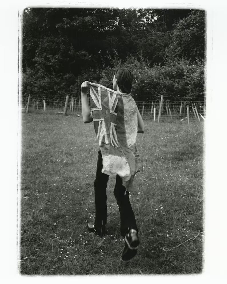 Person walking through a field with a Union Jack flag draped over their shoulders in a black-and-white editorial photograph